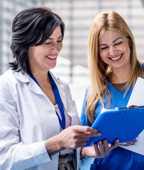 Two smiling healthcare professionals, one in a lab coat, the other in scrubs, reviewing documents in a bright, modern setting.