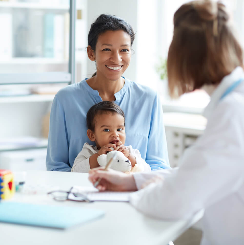 A person holding a baby sits across from another person in a white coat at a desk in a bright, modern room.