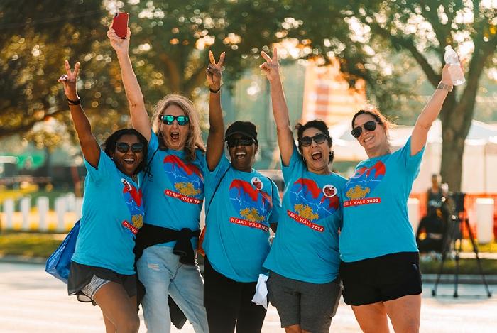 Five people wearing matching blue shirts pose happily, making peace signs outdoors. The scene suggests a casual, joyful event or gathering.
